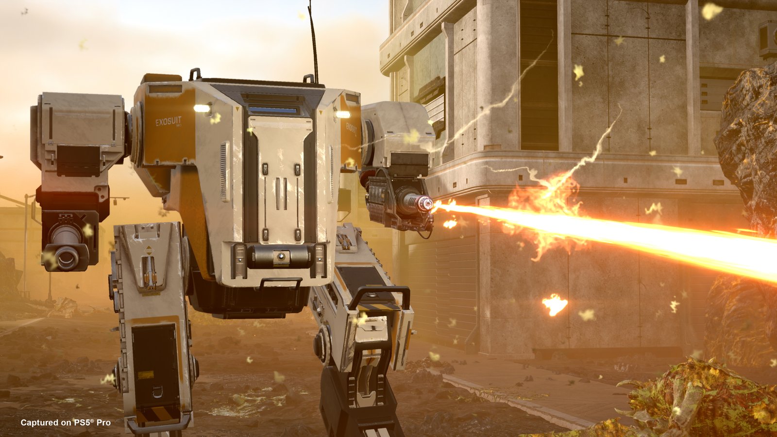 A white and orange exosuit sprays a stream of fire from its arm beside a concrete building