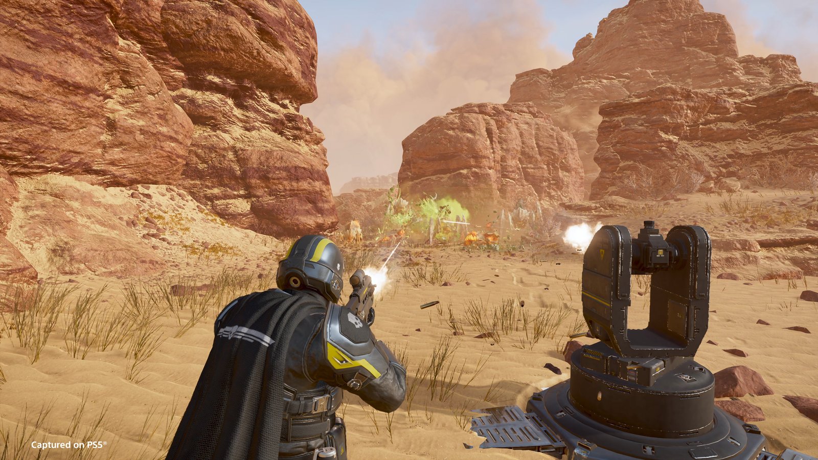 A caped Helldiver fires a rifle beside a sentry turret in a desert canyon as Terminids burst apart in green bile ahead