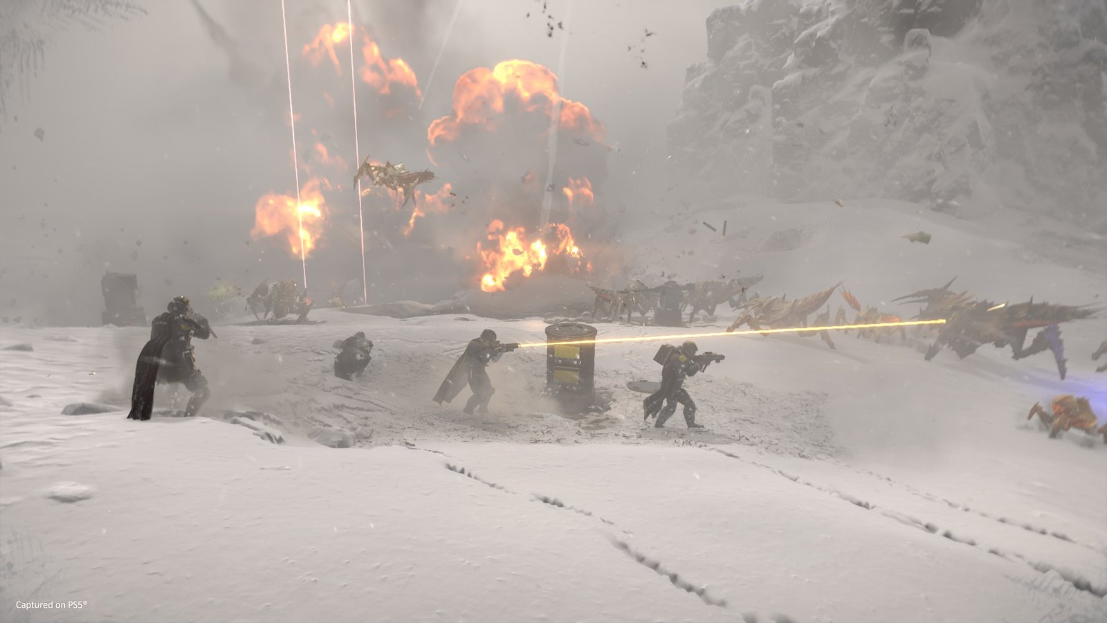 Four Helldivers fire at charging Terminids in the snow beside a supply pod while red stratagem beams and explosions rise behind them