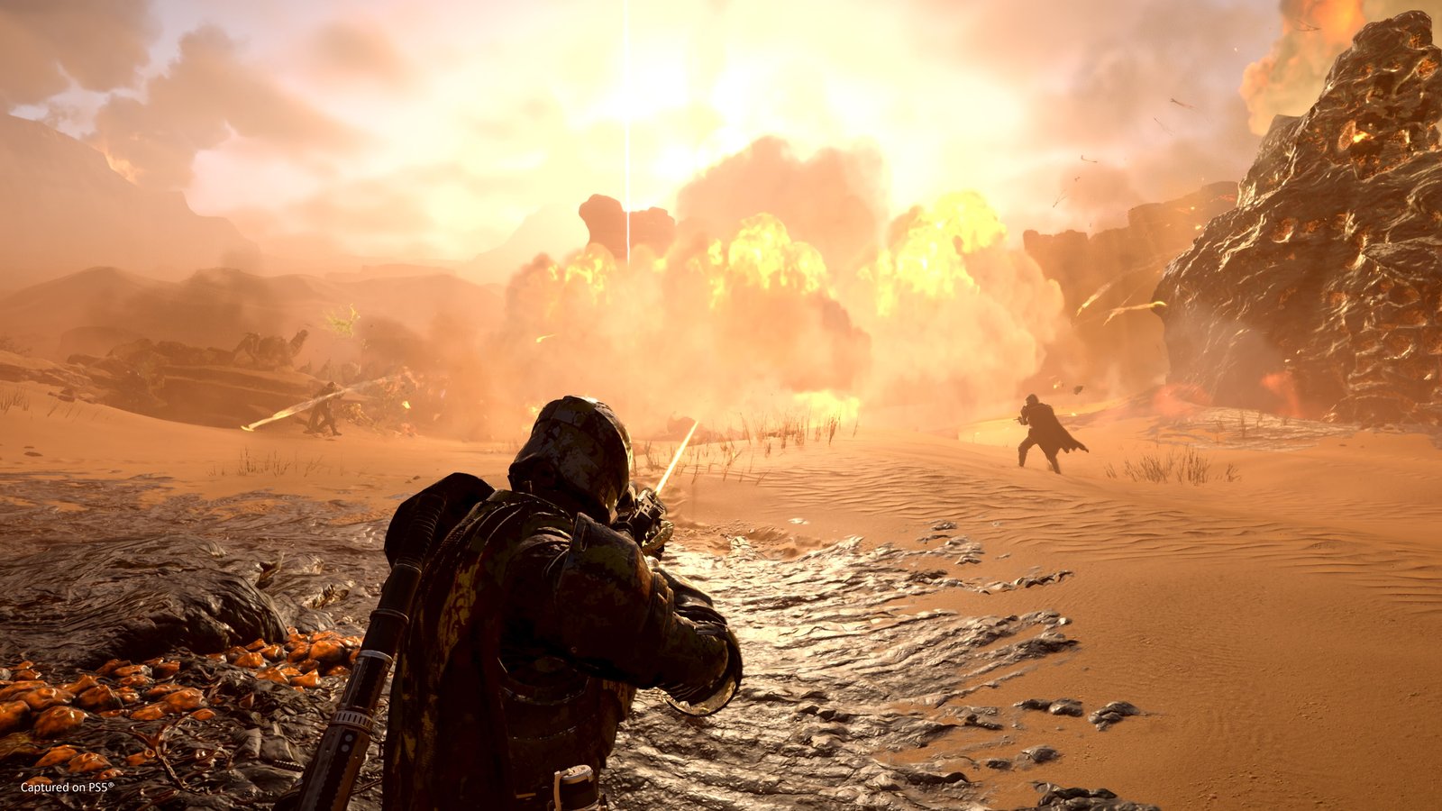 A Helldiver seen from behind fires across a desert as a line of huge explosions erupts ahead and a stratagem beacon's beam rises into the sky